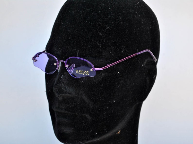 May include: Rimless sunglasses with purple lenses and a thin, purple metal frame. The oval lenses have a solid purple tint. The glasses are displayed on a black mannequin head. The lenses have the text "UV 400 PROTECTION" and "CE".