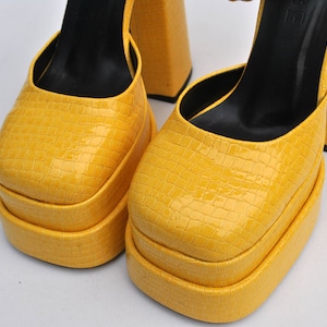 May include: A pair of yellow platform heels with a crocodile-embossed leather upper. The heels are approximately 5 inches tall and have a thick platform.