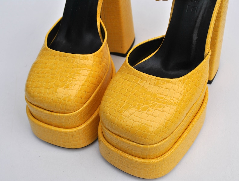 May include: A pair of yellow platform heels with a crocodile-embossed leather upper. The heels are approximately 5 inches tall and have a thick platform.