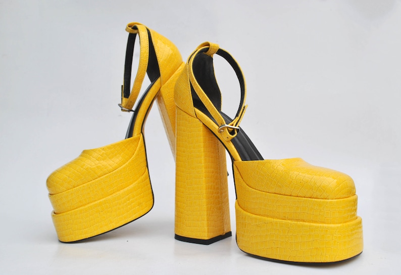 May include: A pair of yellow platform heels with ankle straps. The heels are made of a textured material and have a chunky platform.