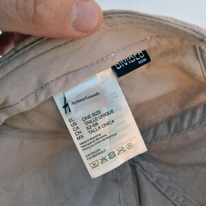 May include: A beige baseball cap with a white label that says "Ariana Grande" and "One Size" in black text. The label also includes size information in different languages. The cap has a black label that says "DIVIDED H&M".
