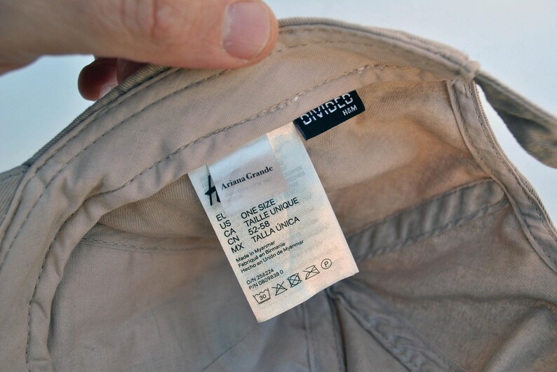 May include: A beige baseball cap with a white label that says "Ariana Grande" and "One Size" in black text. The label also includes size information in different languages. The cap has a black label that says "DIVIDED H&M".