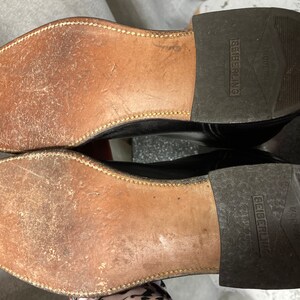 May include: Close-up of the soles of a pair of black leather shoes. The shoes are worn and have a brown leather sole with a stitched edge. The brand name "Seiberling" is printed on the sole of each shoe.