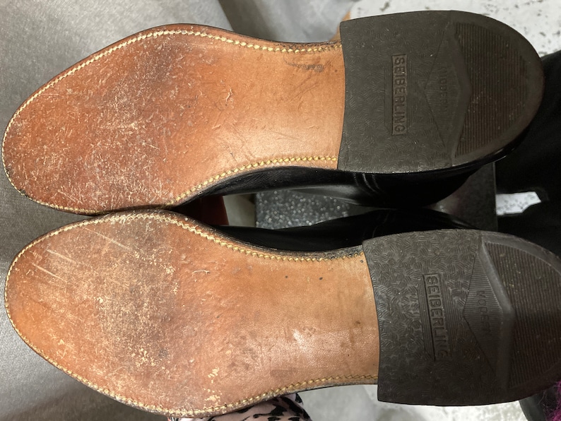 May include: Close-up of the soles of a pair of black leather shoes. The shoes are worn and have a brown leather sole with a stitched edge. The brand name "Seiberling" is printed on the sole of each shoe.