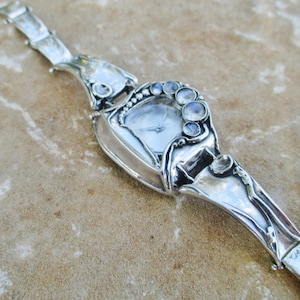 May include: A silver-toned analog wristwatch featuring a polished metal band with organic, flowing lines. The watch face is surrounded by a decorative frame accented with four round, iridescent moonstone cabochons set in silver metal.