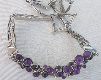 May include: A silver-toned metal bracelet featuring a textured, branch-like structure adorned with numerous round, purple cabochon stones of varying sizes. The piece includes a hinged clasp mechanism and a series of interconnected metal links.