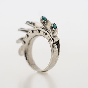 May include: A silver-toned ring featuring a series of articulated, fan-like plates along the outer band. Each plate is accented with a small, round, teal-colored stone. The metal surface has a polished finish with visible rivet-like details.