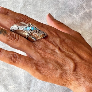 May include: A large, silver-toned statement ring featuring a hammered, organic texture. The centre is adorned with a row of three small, round, iridescent blue opal-like stones, flanked by tiny clear crystals set along the curved, tapered band.