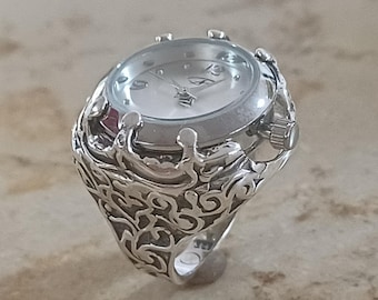 May include: A silver-toned ring featuring an integrated analog watch face with a white dial and silver hands. The band is crafted with an intricate, raised openwork pattern that wraps around the finger, supporting the circular watch housing.