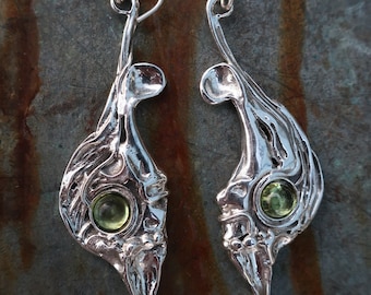 May include: A pair of silver drop earrings featuring an organic, textured metal design with a polished finish. Each earring is set with a single, round, faceted light green gemstone. The earrings hang from simple silver ear wires.