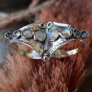 May include: A silver ring featuring a wide, textured, wing-shaped centerpiece set with eight round, iridescent moonstones of varying sizes. The metal surface has a hammered, oxidized finish, and the stones are arranged in a symmetrical row.