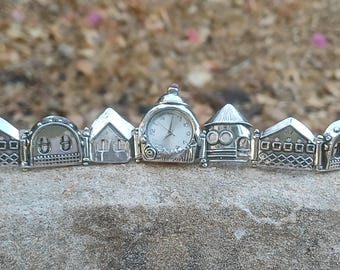 May include: A silver-toned link bracelet featuring a series of miniature house-shaped charms. The central charm is a functional analogue watch with a white face, black hands, and numerals. Each adjacent charm depicts architectural details.