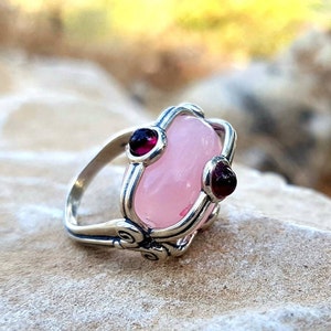 May include: A silver ring featuring a large, oval, pale pink cabochon stone at the center, flanked by two small, round, deep red garnet stones. The silver band displays intricate metalwork with swirling patterns around the stone settings.
