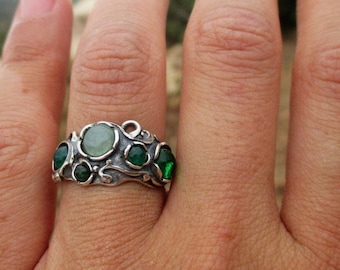 May include: A silver ring featuring a textured, organic metal band adorned with one central pale green round stone and four smaller, vibrant dark green round stones set within the swirling, raised metalwork of the band's surface.