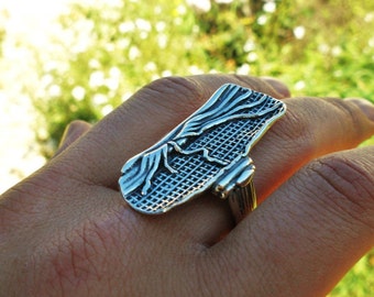 May include: A silver-toned ring featuring an elongated, rectangular face with a textured surface. The design includes raised, flowing lines that resemble tree branches or roots layered over a grid-like pattern, finished with a polished metal band.