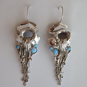 Könnte beinhalten: Ein Paar silberfarbene Ohrhänger mit einer fließenden, organischen Form, die an eine Qualle erinnert. Die Metalloberfläche ist poliert und reflektierend und mit kleinen, runden, hellblauen Cabochon-Steinen an den Seiten verziert.