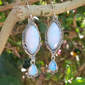 May include: A pair of dangle earrings featuring faceted, marquise-shaped opalescent stones set in oxidized silver frames. Each earring includes a smaller, teardrop-shaped opalescent stone suspended from the base, attached by a silver hook wire.