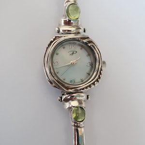 May include: Silver wristwatch with a round face and a mother-of-pearl dial. The watch features a silver band with green gemstone accents. The watch face has the numbers 12, 3, 6, and 9 marked with silver hands.