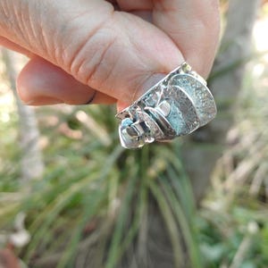 Puede incluir: Un anillo de plata con una banda ancha y texturizada con un diseño abstracto en capas. La superficie del metal tiene un acabado rugoso y martillado con bordes pulidos, creando una apariencia multidimensional en la parte frontal.