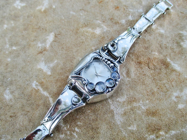 May include: A silver-toned wristwatch featuring a polished, organic-shaped metal band and case. The watch face is round with silver hands, and the bezel is accented with a cluster of five round, translucent, iridescent cabochon gemstones.