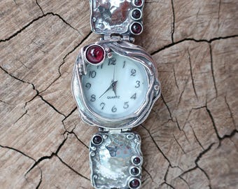 May include: A silver watch with a white face and black hands. The watch has a unique design with a textured band and red gemstones. The watch is on a wooden surface.