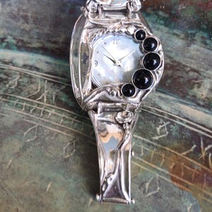 May include: A silver-toned watch with an organic, fluid metal frame and a mother-of-pearl dial. The watch features a series of four round, polished black stones arranged in a crescent shape along the right side of the watch face.
