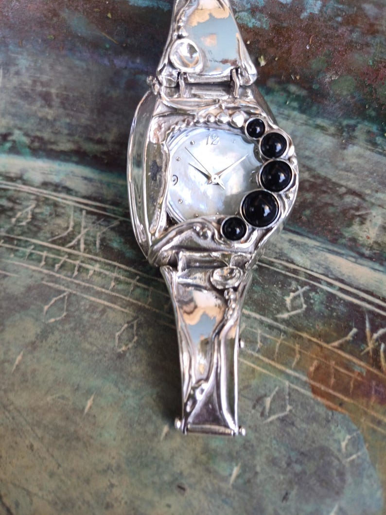 May include: A silver-toned watch with an organic, fluid metal frame and a mother-of-pearl dial. The watch features a series of four round, polished black stones arranged in a crescent shape along the right side of the watch face.