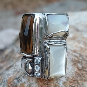 Könnte beinhalten: Ein Silberring mit einem rechteckigen, facettierten braunen Edelstein, zwei glatten weißen rechteckigen Einlagen und einer vertikalen Reihe aus drei kleinen, klaren runden Steinen in einer texturierten Metallfassung mit oxidiertem Finish.