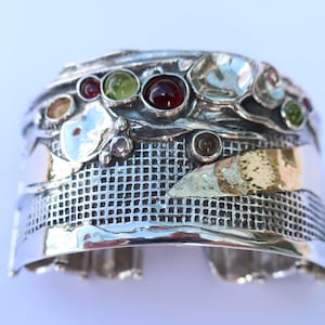 Op de afbeelding: Een brede zilveren manchetarmband met een gestructureerd rasterpatroon in het midden, geaccentueerd met een gepolijste goudkleurige metalen strip. De bovenrand is versierd met diverse ronde, veelkleurige edelstenen in rood, groen en amber.