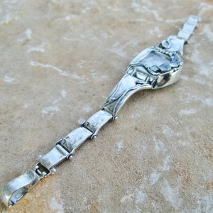 May include: A silver-toned metal bracelet featuring a series of rectangular links connected by small hinges. The central focal point is a larger, curved silver-toned element adorned with three small, round, iridescent cabochon stones.