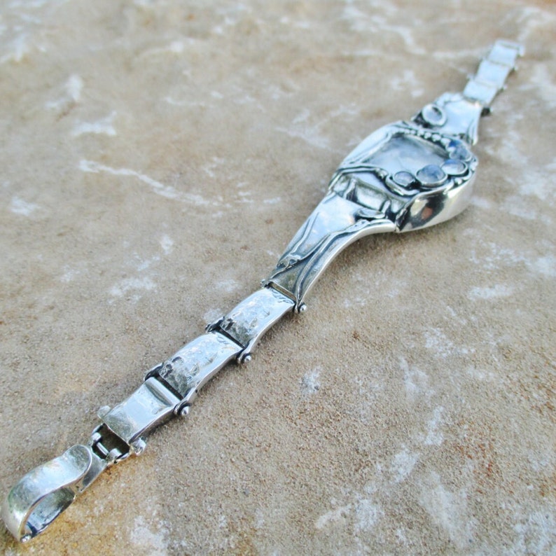 May include: A silver-toned metal bracelet featuring a series of rectangular links connected by small hinges. The central focal point is a larger, curved silver-toned element adorned with three small, round, iridescent cabochon stones.