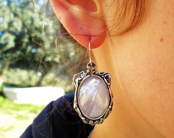 May include: An oval-shaped earring featuring a central iridescent mother-of-pearl cabochon set within a detailed, oxidized silver frame. The metalwork displays intricate scrollwork and organic textures, suspended from a simple silver hook wire.