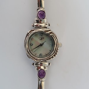 May include: A silver watch with a mother-of-pearl face and purple gemstone accents. The watch has a unique, twisted band.