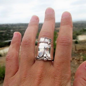 May include: A wide sterling silver ring featuring a multi-layered, textured band design with a polished finish. The ring includes small, raised circular accents and a single round, smooth stone set into the metal surface, creating a layered appearance.