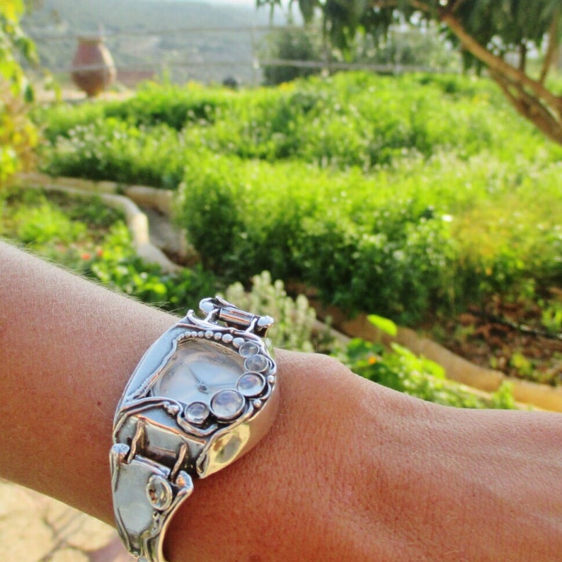 May include: A silver-toned wristwatch featuring a uniquely shaped case with a curved, organic design. The watch face is surrounded by a series of small, clear, round stones set along the side, and it is secured with a matching silver-toned metal band.