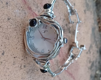 May include: A silver-tone wristwatch featuring a round white dial with silver numerals at the 3, 6, 9, and 12 o'clock positions. The watch case is surrounded by a textured silver frame accented with two black cabochon stones and a link bracelet.