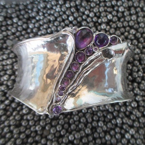 Puede incluir: Un brazalete ancho de plata con un acabado martillado y reflectante. La pulsera presenta una hilera diagonal de piedras preciosas de amatista púrpura, redondas y en forma de pera, que crean un contraste sobre la superficie metálica pulida.