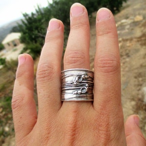 Op de afbeelding: Een brede zilveren ring met een getextureerd oppervlak van horizontale lijnen en kleine verhoogde cirkelvormige accenten. Het metaal heeft een gepolijste afwerking met donkere verdiepte gebieden die de lineaire details benadrukken.