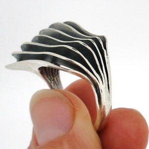 May include: A silver ring featuring a series of five stacked, curved, and layered metal plates that fan out from the band. The metal has a polished finish on the edges and a darkened, oxidized appearance within the recessed areas between the layers.