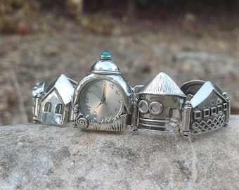 May include: A silver watch bracelet with a central clock face and a turquoise accent. The bracelet is decorated with miniature house and building designs. The watch has a silver face with black numerals and hands.