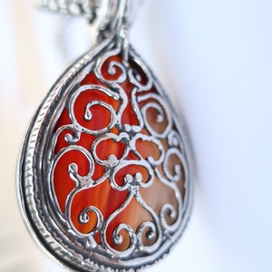 Op de afbeelding: Een druppelvormige hanger met een levendige oranje, gestreepte steen in een kooi van zilverkleurig metaal met ingewikkeld filigraanwerk. De hanger hangt aan een bijpassende zilverkleurige ketting met gedetailleerde krulpatronen.