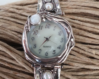 May include: A silver-toned quartz wristwatch featuring a white dial with black numerals and hands. The watch case and band have a textured, organic metal finish and are accented with small, round white pearls embedded in the metal links.