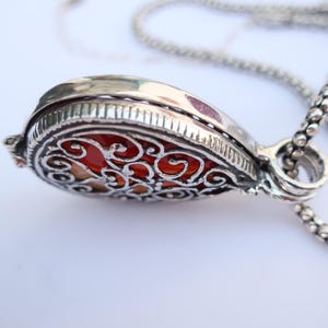 Op de afbeelding: Een zilverkleurige hangerketting met een sierlijk filigraanontwerp. De druppelvormige hanger onthult een rood-oranje steen door het opengewerkte ontwerp. De ketting heeft een delicate, kralenachtige stijl.