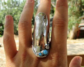 May include: A long, silver-toned statement ring featuring a smooth, reflective, elongated surface. The base of the ring is accented with two round, bright blue cabochon stones and a single, smaller, clear, faceted round stone set in silver metal.