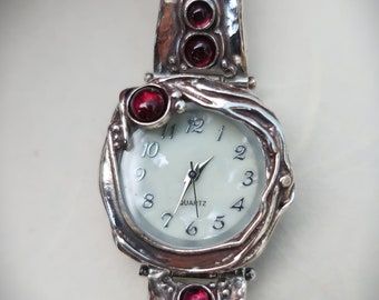 Puede incluir: Un reloj de pulsera plateado con una esfera blanca y marcadores de hora romanos negros. El reloj tiene un diseño único y orgánico con acentos de piedras preciosas rojas en la banda y la esfera.