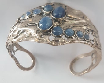 May include: A silver-toned cuff bracelet featuring an organic, textured metal surface with an openwork design. The bracelet is adorned with multiple smooth, round, blue cabochon stones of varying sizes arranged in a cluster across the front.