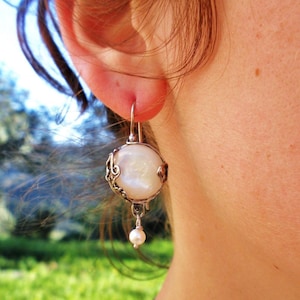 May include: A silver dangle earring featuring a large, round, iridescent white pearl encased in an ornate, swirling silver wire frame, with a smaller, round white pearl suspended from the bottom by a thin silver wire hook.
