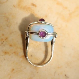 Puede incluir: Un anillo giratorio de plata con una piedra rectangular blanca translúcida similar a un ópalo en un marco de metal. El centro de la piedra está acentuado con una pequeña gema cabujón redonda de color púrpura y otra piedra púrpura en la parte superior.