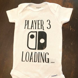 May include: White baby onesie with the text "PLAYER 3 LOADING" in black. The design includes a stylized graphic resembling a game controller. A cute and playful garment for a new baby.