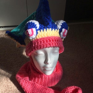 May include: A pink crocheted hat and scarf set. The hat has a blue, yellow, and green crocheted design on top. The hat and scarf are both made with a textured stitch.